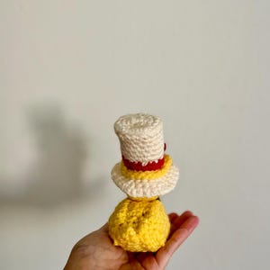 May include: A crocheted yellow and white doll wearing a red and white hat. The doll is being held in a hand.