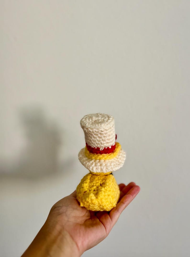 May include: A crocheted yellow and white doll wearing a red and white hat. The doll is being held in a hand.