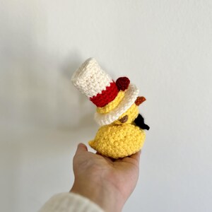 May include: A crocheted yellow chick wearing a red, white, and yellow hat with a red pom-pom.