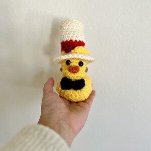 May include: A yellow crocheted chick wearing a white and red top hat with a black bow tie.