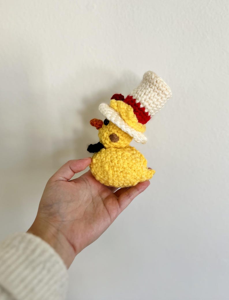 May include: A yellow crocheted duck wearing a white and red top hat. The duck is being held in a person's hand.