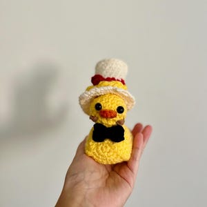 May include: A yellow crocheted duck wearing a white and red top hat with a black bow tie.