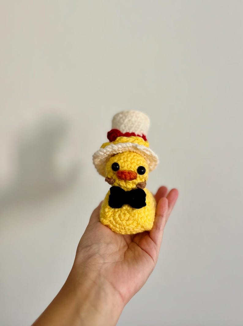 May include: A yellow crocheted duck wearing a white and red top hat with a black bow tie.