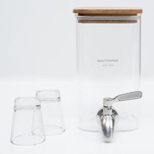 May include: A clear glass dispenser with a wooden lid and a stainless steel spigot. The dispenser is labeled "MOUTHWASH 1500ml / 42.3 fl oz". Two clear glass cups are also included.