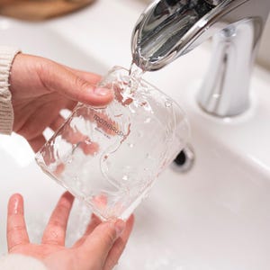 May include: A clear glass toothbrush holder with the text "TOOTHBRUSH 500ml / 16.9 fl oz" being filled with water from a chrome faucet.