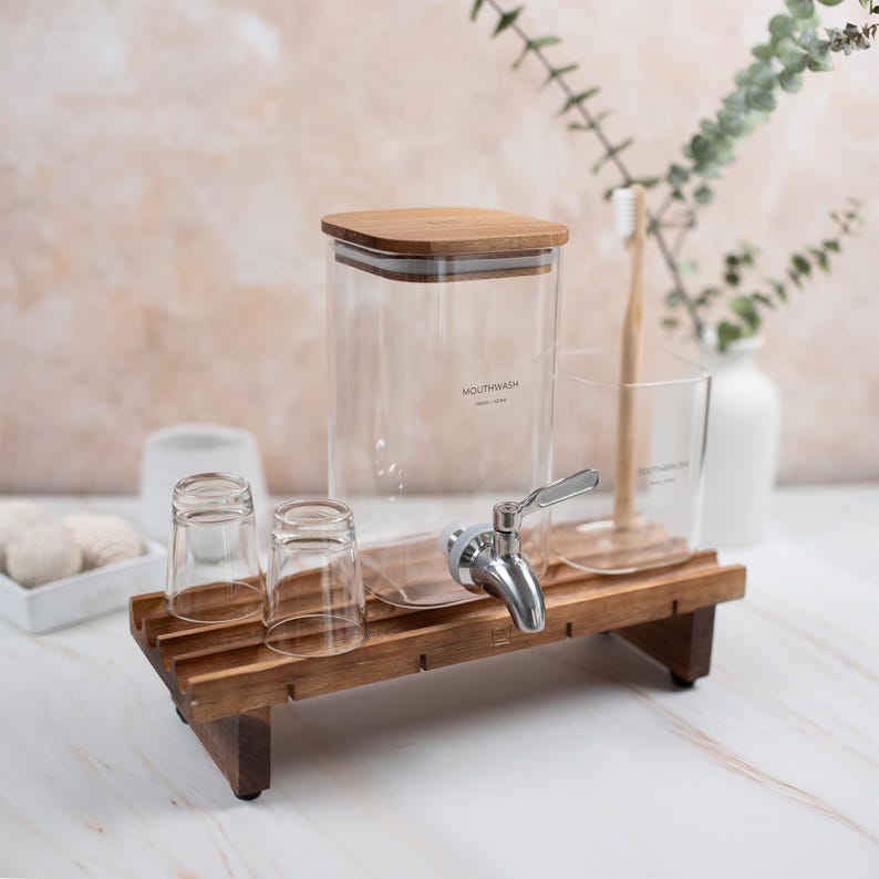 May include: A wooden stand with a glass dispenser labeled "Mouthwash" and two small glasses. The dispenser has a silver tap and a wooden lid.