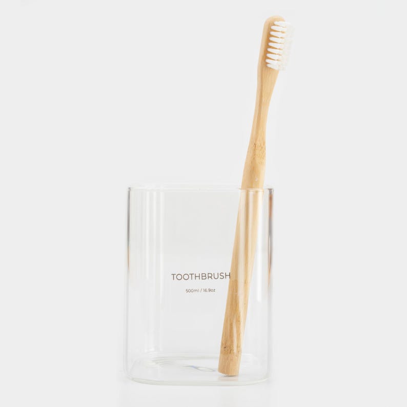 May include: A clear glass toothbrush holder with a bamboo toothbrush inside. The holder has the text "TOOTHBRUSH 100ml / 16.9oz" printed on the side.