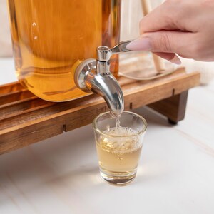 May include: A glass dispenser with a silver spigot pouring a clear liquid into a glass shot glass. The dispenser is sitting on a wooden stand.