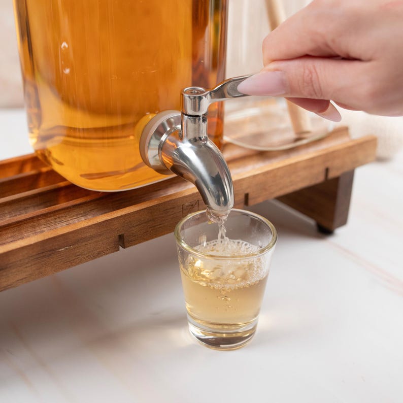 May include: A glass dispenser with a silver spigot pouring a clear liquid into a glass shot glass. The dispenser is sitting on a wooden stand.