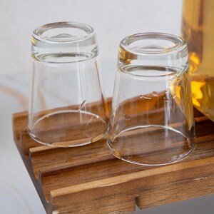 May include: Two clear glass shot glasses on a wooden tray. The glasses are empty and have a slight sheen from the light reflecting off of them.