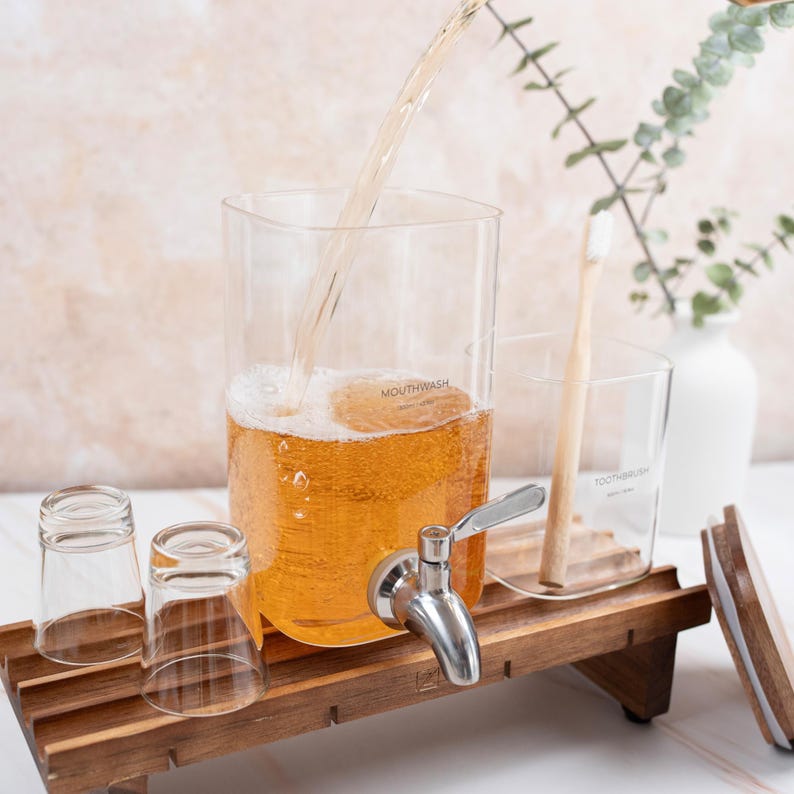 May include: A wooden countertop with a glass dispenser filled with amber liquid labeled "MOUTHWASH". Two clear glass cups and a glass jar labeled "TOOTHBRUSH" are on the countertop. A wooden tray with a faucet is under the dispenser.
