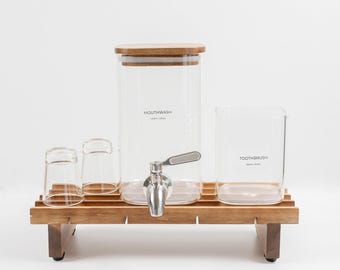 May include: A clear glass mouthwash dispenser with a wooden lid and silver spigot, a clear glass toothbrush holder, and two small clear glass cups, all arranged on a slatted wooden tray. The dispenser is labeled MOUTHWASH and the holder TOOTHBRUSH.