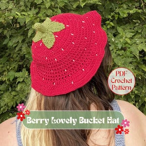 May include: A red crochet bucket hat, designed to look like a strawberry, with white seed details. It has a green crocheted stem and leaf. The image includes the text "Berry Lovely Bucket Hat" and "PDF Crochet Pattern".