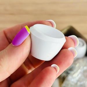 May include: A close-up of a white ceramic candle holder. The holder is round and has a smooth, glossy finish. It is being held in a hand with pink and yellow nail polish.
