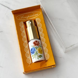 May include: A small perfume atomiser with a white ceramic base featuring a painted floral design of red, blue, and yellow flowers. The atomiser has a gold-coloured metal spray nozzle and is housed in an orange cardboard box labelled Pompadour.