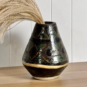May include: A tapered ceramic vase with a glossy dark green and brown glaze featuring a geometric pattern of triangles and diamonds. The vase has a wide base and a narrow neck, and it contains a large, feathery bunch of dried beige pampas grass.