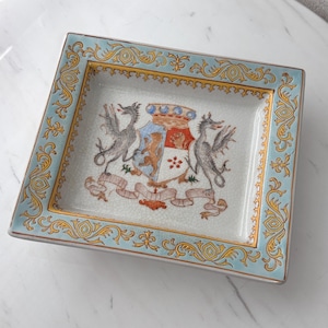 May include: Square ceramic tray with a light blue border and gold filigree design. The centre features a coat of arms with two grey dragons, a shield, and a banner. The tray is likely for decorative use.
