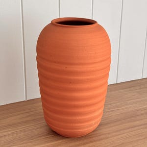 May include: A terracotta-coloured, ribbed vase with a rounded body and a wide opening. The vase has a textured surface and sits on a wooden surface. The background is a white wall with vertical panelling.