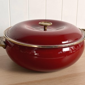 May include: A deep, round, glossy dark red cooking pot with a matching lid. The pot features two curved brass-toned metal handles on the sides and a circular brass-toned knob on the centre of the lid, with a thin brass-toned rim around the edge.