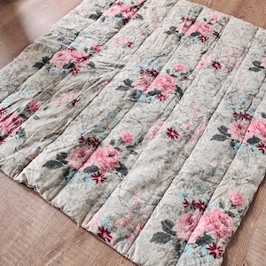 May include: A quilted blanket with a floral design. The blanket has a grey background with pink roses, blue accents, and dark green leaves. The rectangular blanket has vertical stitching.