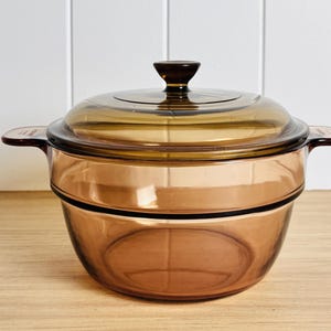 May include: An amber glass casserole dish with a lid. The dish has two side handles and a dark brown knob on the lid. The cookware is designed for oven use. The dish is suitable for cooking and serving.