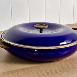 May include: A round, deep blue enamelled cooking pan with a matching lid. The pan features two curved brass-toned side handles and a central brass-toned knob on the lid. The rim of the pan and the lid edge are finished with a metallic brass-toned trim.