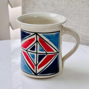 May include: A ceramic mug with a geometric design. The mug has a cream-colored base and handle, with a pattern of red, blue, and navy blue shapes outlined in black. The mug is designed for hot drinks.