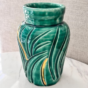 May include: A glossy emerald green ceramic vase featuring a relief pattern of elongated, curved leaves. The surface has a smooth, reflective finish with thin, vertical gold-painted accents highlighting the contours of the sculpted leaf design.