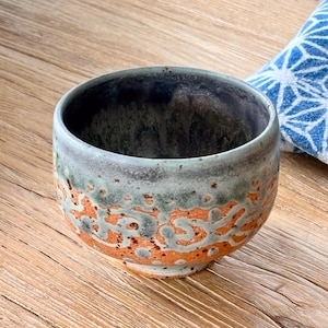 May include: A ceramic tea bowl featuring a rounded shape with a wide opening. The exterior displays a textured surface with a blend of earthy orange and muted green glaze, while the interior is finished in a deep, dark charcoal tone.