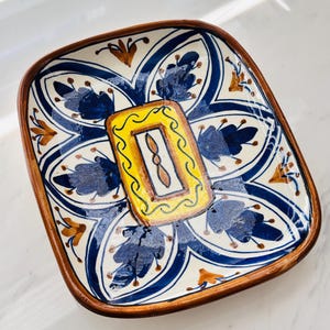 May include: A square ceramic dish with rounded corners, featuring a hand-painted floral pattern in deep blue and golden brown on a white background. The centre displays a bright yellow rectangular border surrounding a small brown leaf motif.