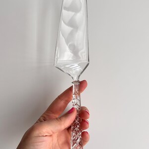 May include: A clear glass cake server with a diamond-patterned handle. The server has a wide, curved blade and a decorative, textured handle. The server is designed for serving cakes and other desserts.