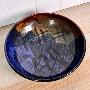 Peut inclure: Un bol en céramique rond présentant un vernis brillant avec un dégradé de couleurs passant du bleu profond à l'ambre chaud et au brun. La surface intérieure affiche un motif texturé et tacheté avec des marques de pinceau sombres.