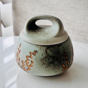 May include: A small, round ceramic jar with a lid and handle. The jar is a muted green colour with brown floral accents. The lid has a curved handle. The jar is on a white surface.