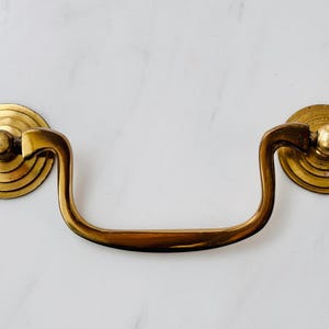 May include: A brass drawer pull featuring a curved, swinging handle attached to two circular, tiered backplates. The metal has a polished, reflective finish with subtle signs of age and wear, showcasing a classic, functional hardware design.