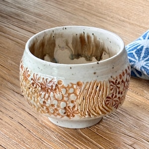 May include: A ceramic tea bowl with a speckled off-white glaze and a textured exterior featuring carved floral patterns and vertical lines in shades of brown. The interior shows brown glaze drips running down from the rim onto the cream base.