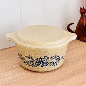 May include: A vintage beige casserole dish with a matching lid. The dish has a speckled finish and a blue floral pattern around the base. The handles are curved. A decorative wooden cat figurine is in the background. The dish is likely around 20 cm in diameter.