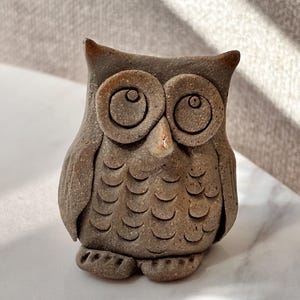 May include: A small, hand-sculpted clay owl figurine in a muted brown, earthy tone. The owl features large, circular eyes with defined pupils, a pointed beak, and textured, overlapping semi-circular patterns representing feathers on its chest.