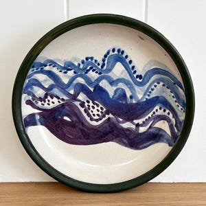 May include: A round ceramic plate with a dark green rim and an off-white base. The centre features hand-painted abstract wavy lines in shades of blue and deep purple, accented with small dots along the curves, creating a layered wave pattern.