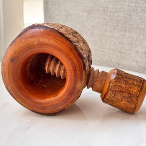 May include: A wooden nutcracker featuring a circular base with natural bark edges and a smooth, polished interior. A threaded screw mechanism with a matching bark-textured handle extends from the side, designed for manual operation to crack nuts.