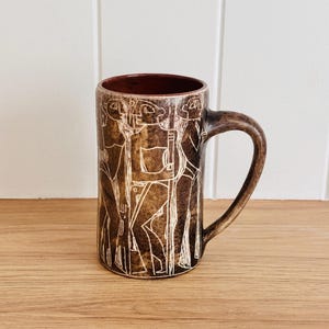 May include: A brown ceramic mug with a white design of stylized figures. The mug has a rounded handle.