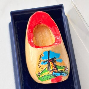 May include: A miniature wooden clog with a natural light wood finish and a bright red interior rim. The exterior features a colourful painted illustration of a windmill and a landscape with the word HOLLAND printed in black vertical text.