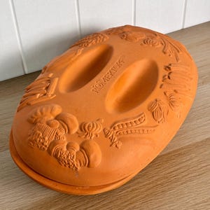 May include: An oval-shaped terracotta Romertopf clay baker with a lid featuring two recessed handles and embossed vegetable motifs including mushrooms, onions, and peas. The natural orange-brown clay surface has a matte finish and texture.