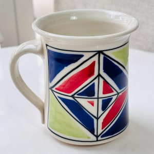 May include: A ceramic mug with a geometric pattern. The mug has a white rim and handle, and the body is decorated with blue, red, and green geometric shapes outlined in black. Suitable for hot or cold drinks.