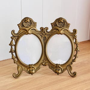 May include: A vintage brass double photo frame with ornate detailing. The frame has two oval openings for photos, with decorative flourishes and a sturdy base. The frame is a warm gold colour and stands upright on a wooden surface.