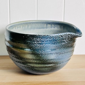 May include: A ceramic mixing bowl with a deep blue and green glaze featuring a speckled white texture. The bowl has a rounded shape with horizontal ridges along the exterior and a small integrated pouring spout on the upper right rim.