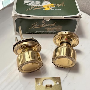 May include: Two polished brass door knobs with a circular rope-textured trim, a rectangular brass strike plate, and a green and white Gainsborough Stronghold Series box with the text "THE KEY TO HOME SECURITY" and a yellow Hudson price sticker.