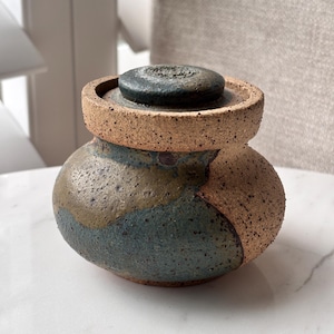 May include: A small, round, lidded ceramic jar with a textured, speckled surface. The body features a combination of earthy brown, muted green, and dark blue glazes, while the lid is a dark, matte charcoal colour with a rough, granular finish.