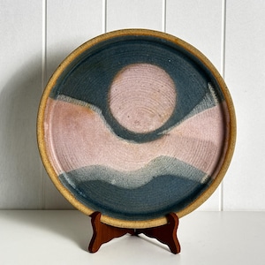 May include: A round ceramic plate featuring a textured surface with a colour palette of muted pink and dark blue. The design includes a circular pink centre surrounded by dark blue waves. The rim of the plate is a natural tan, earthy clay colour.