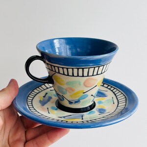 May include: A ceramic teacup and saucer set. The cup has a blue rim and a black handle. The cup and saucer are decorated with colourful abstract shapes. The saucer has a blue rim and a white centre.