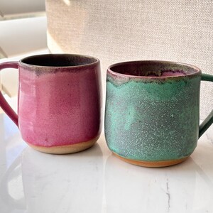 May include: Two ceramic mugs with rounded bodies and handles. The left mug features a vibrant pink glaze with a dark rim, while the right mug has a textured green glaze with a dark rim. Both mugs have an exposed, unglazed tan clay base.